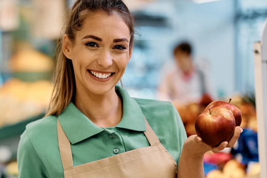 Lächelnde Supermarktmitarbeiterin im grünen Polo hält zwei rote Äpfel in der Obstabteilung.