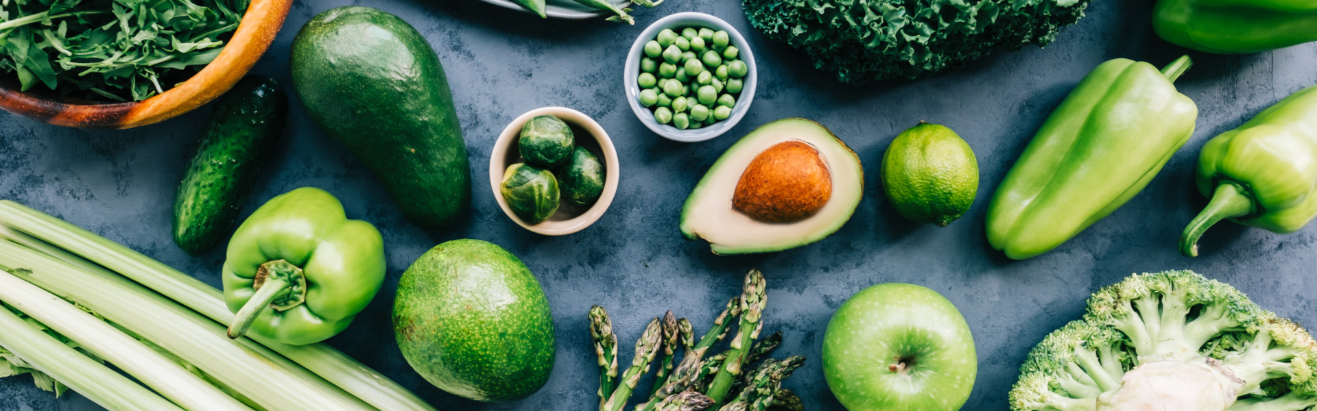 Verschiedene grüne Obst- und Gemüsesorten wie Avocado, Erbsen, Rosenkohl, Spargel und Apfel auf dunkler Oberfläche.