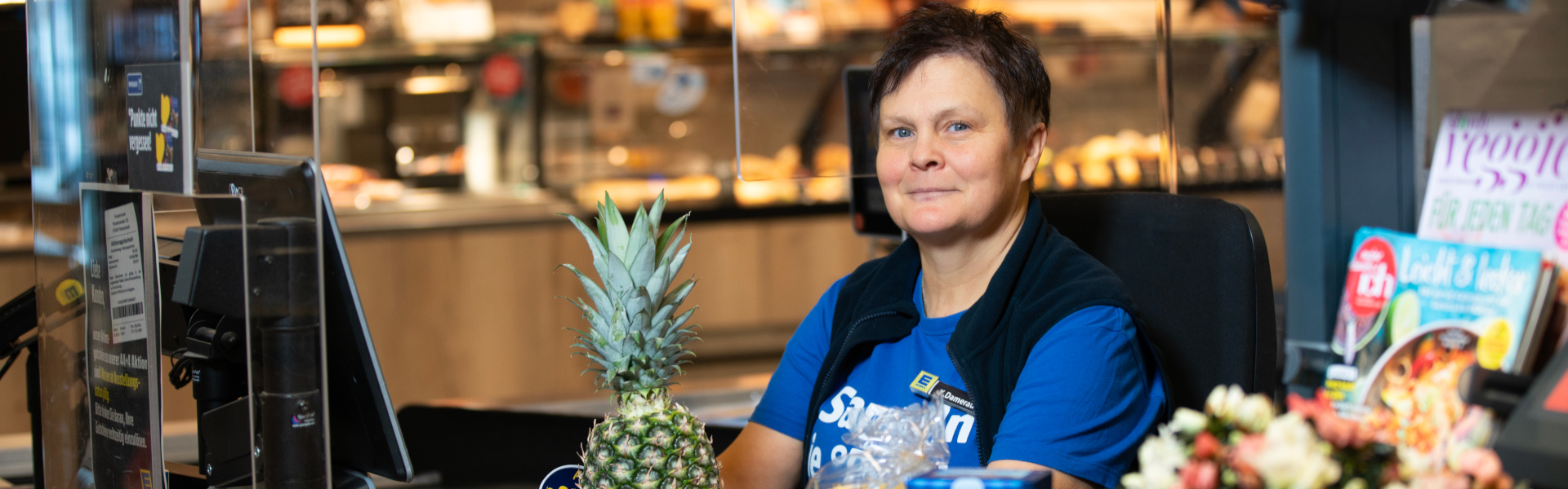 Mitarbeiterin an der Kasse in einem blauen Payback Shirt kassiert und lächelt in die Kamera